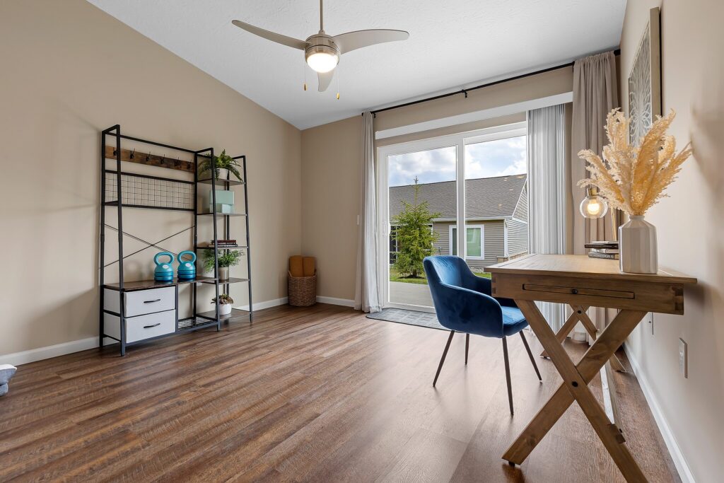 A home office with a wooden desk, blue chair, shelving unit, and decor, featuring large glass doors that open to a view of a grassy yard and neighboring house.
