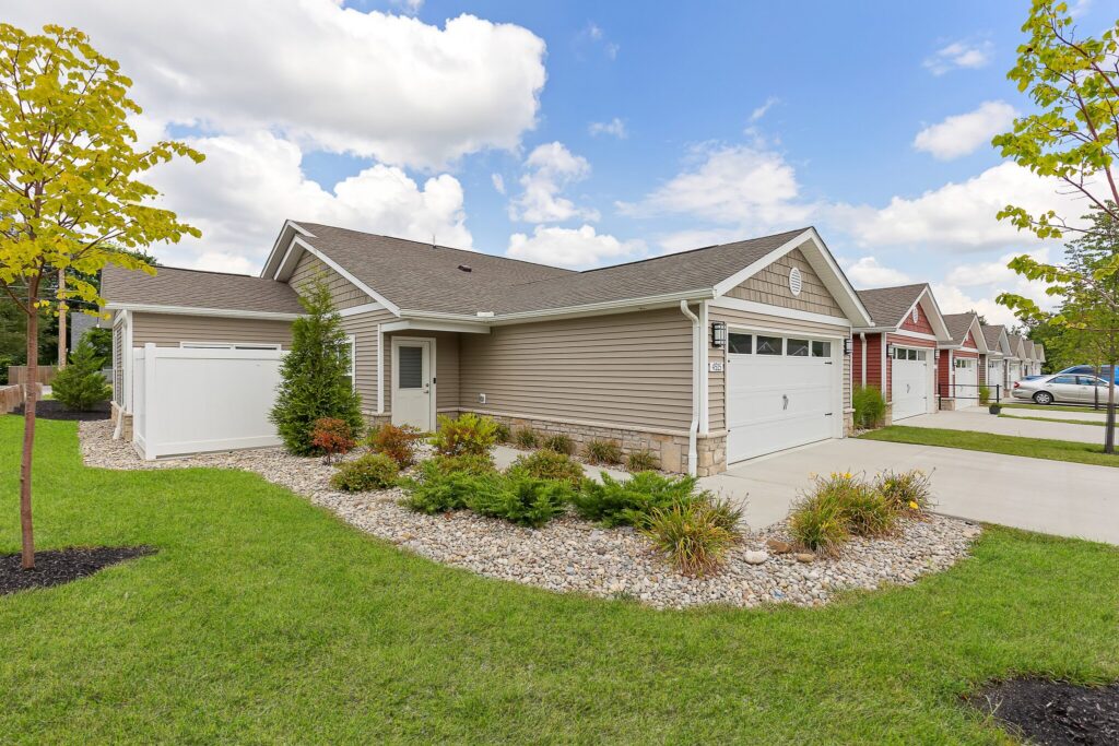 Single-story suburban house with beige siding, a two-car garage, manicured lawn, and landscaped front yard with shrubs and rocks, under a partly cloudy sky.