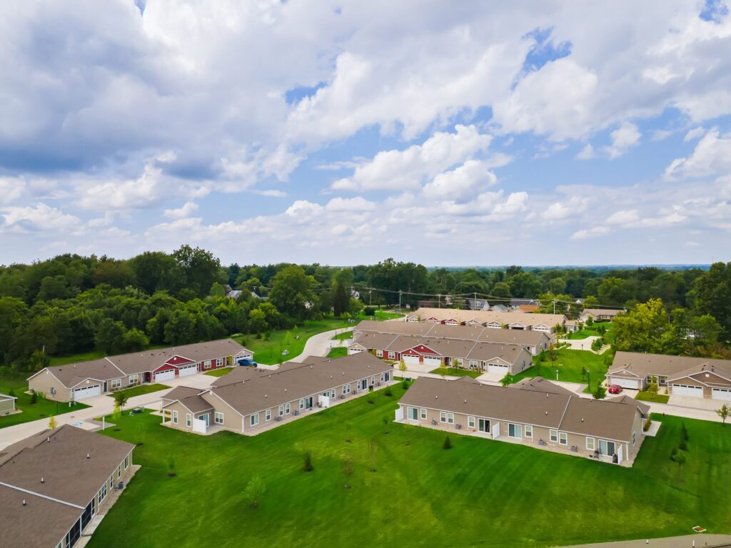 Aerial view of a suburban neighborhood with single-story houses, green lawns, trees, and a partially cloudy sky.