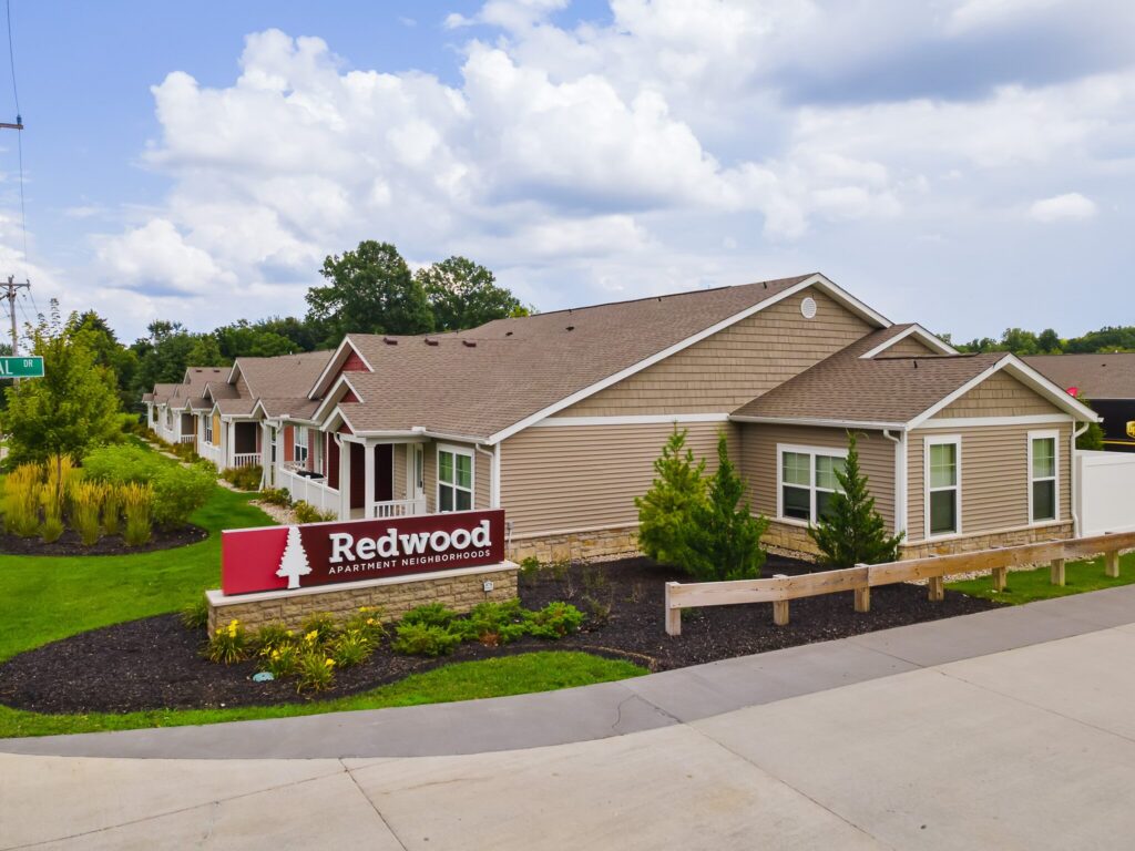Single-story apartment buildings with beige siding, white trim, and gabled roofs; a sign in front reads "Redwood Apartment Neighborhoods.