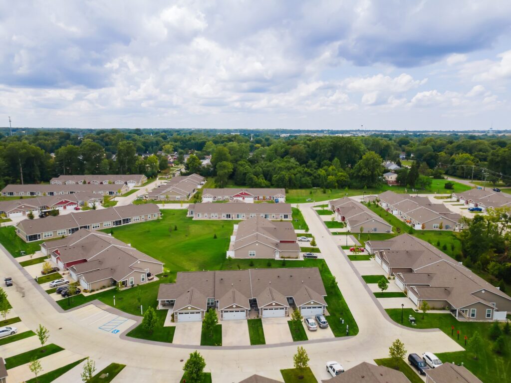 Aerial view of a suburban neighborhood with rows of single-family homes, green lawns, driveways, and surrounding trees under a partly cloudy sky.