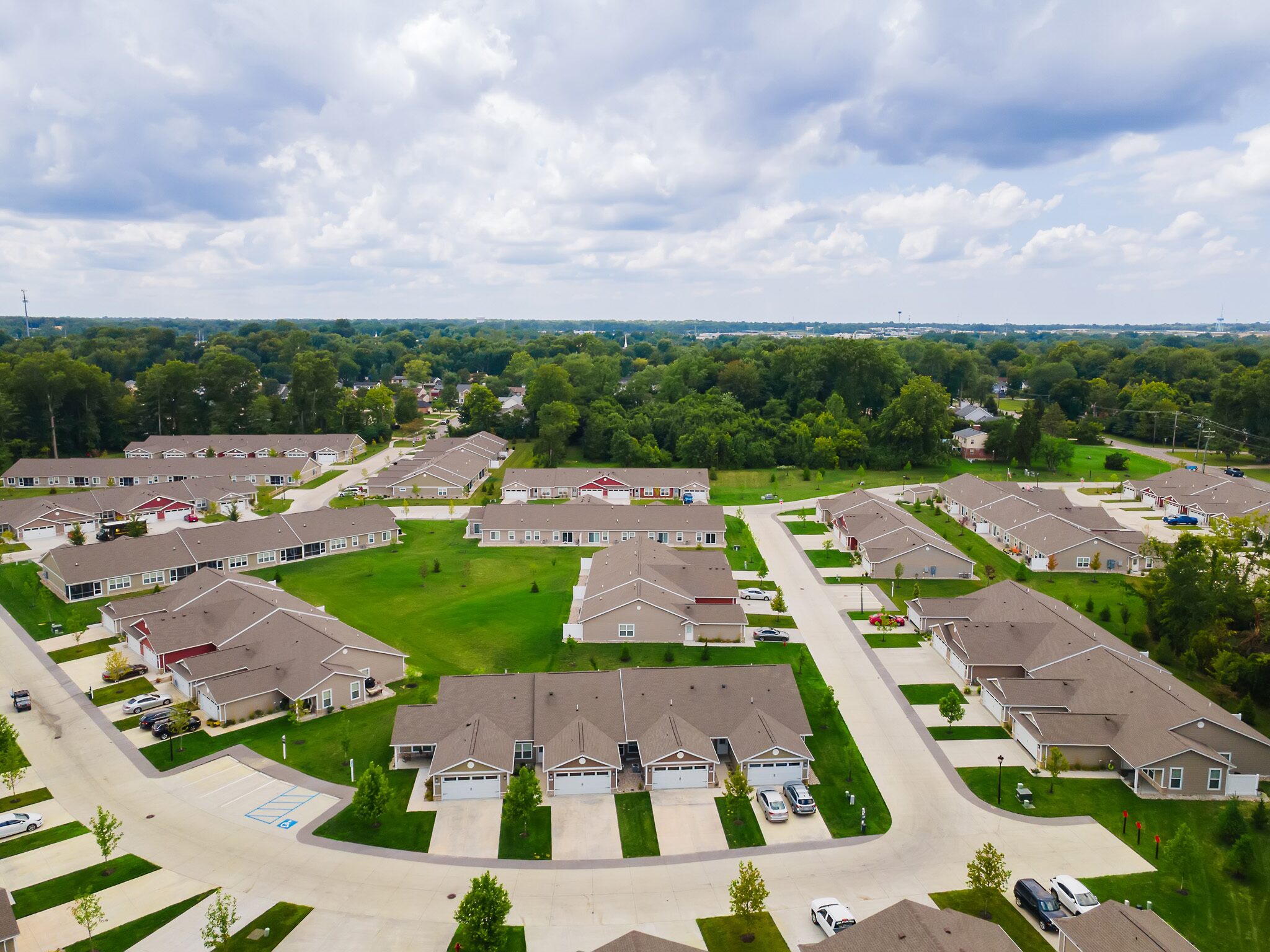 Aerial view of a suburban neighborhood with rows of single-family homes, green lawns, driveways, and surrounding trees under a partly cloudy sky.