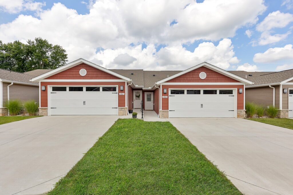 Duplex with red siding, two white garage doors, and concrete driveways, separated by a grass lawn under a partly cloudy sky.