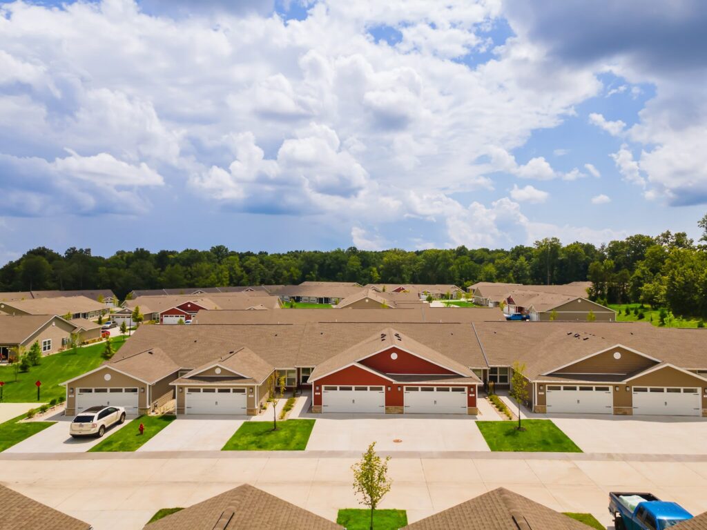 A row of single-story suburban houses with attached garages, neatly trimmed lawns, cars parked in driveways, and wooded area in the background under a partly cloudy sky.