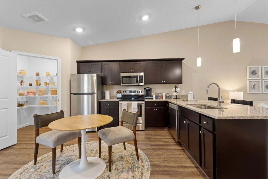Modern kitchen with dark wood cabinets, stainless steel appliances, granite countertops, round dining table with two chairs, and pantry visible through an open door.