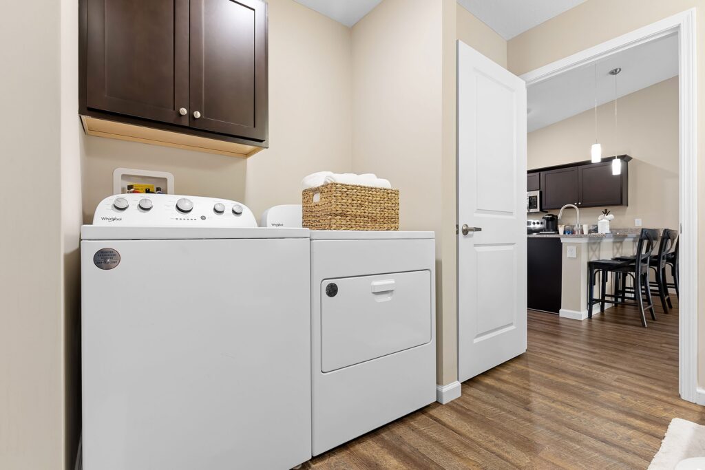 Laundry room with white Whirlpool washer and dryer, a brown cabinet above, and a wicker basket on the dryer; open door leads to kitchen and dining area with wood flooring.