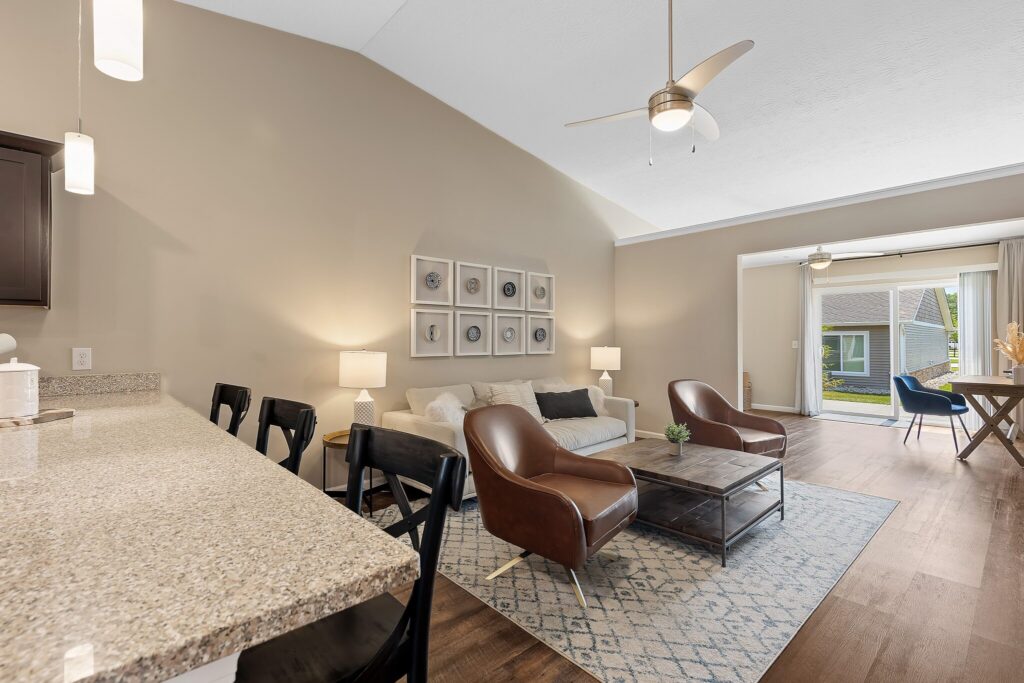 Open-concept living room with a sofa, two leather chairs, a coffee table, and large window doors leading to a patio; kitchen counter with barstools in the foreground.