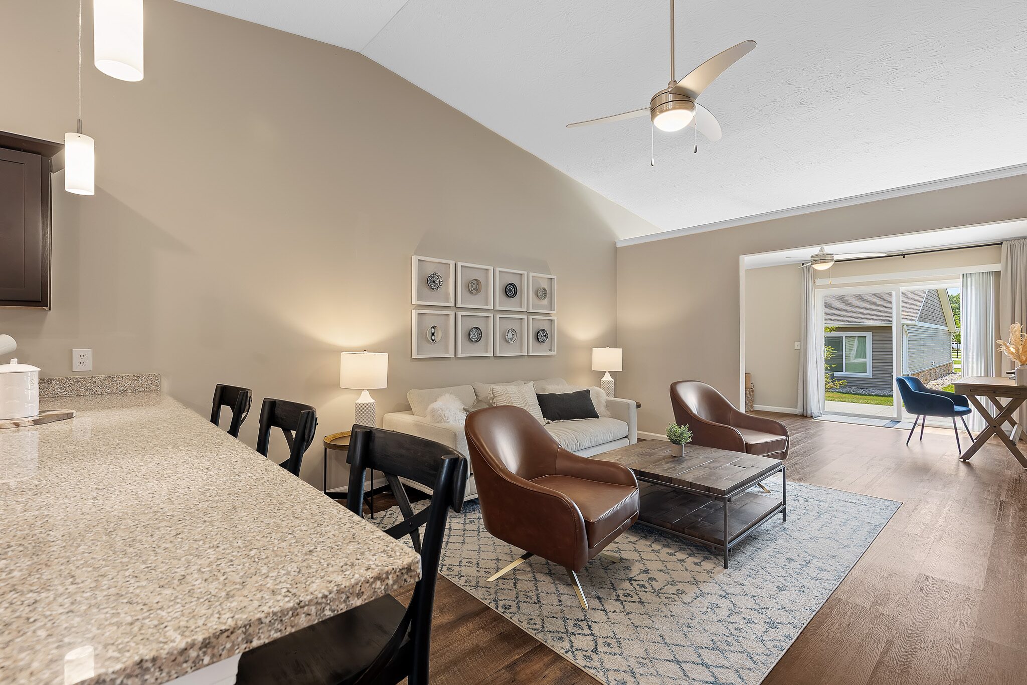 Open-concept living room with a sofa, two leather chairs, a coffee table, and large window doors leading to a patio; kitchen counter with barstools in the foreground.