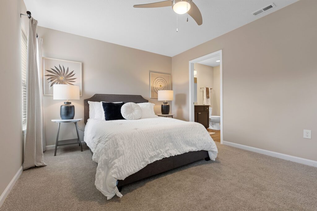 A bedroom with a neatly made bed, two dark bedside tables with lamps, neutral walls, beige carpet, ceiling fan, and a view into an adjoining bathroom.