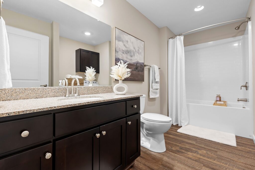 Modern bathroom with dark wood cabinets, granite countertop, wall art, white fixtures, a bathtub with a shower curtain, and wood-look flooring.