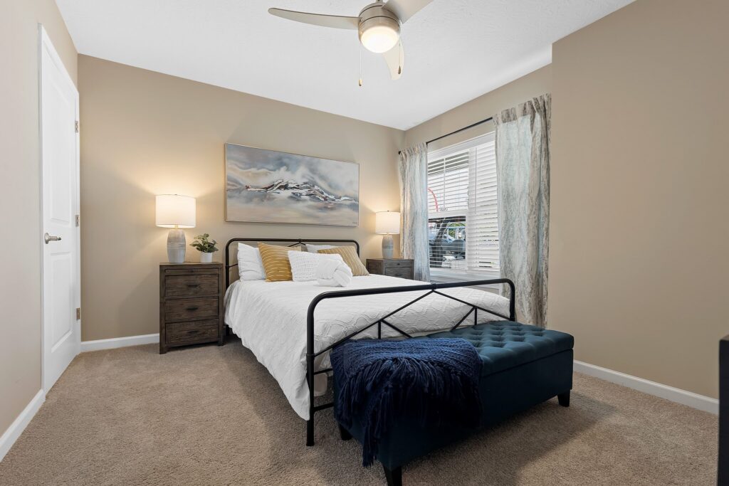 A bedroom with a bed, white bedding, two bedside tables with lamps, a wall painting, a window with sheer curtains, and a blue bench at the foot of the bed.