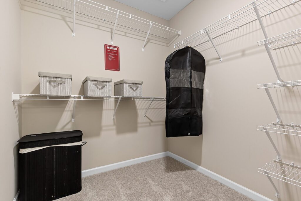 A walk-in closet with white wire shelving, storage boxes, a black hamper, and a garment bag hanging on the wall. Carpeted floor and beige walls.