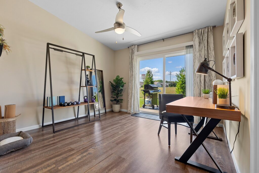 Modern home office with desk, chair, bookshelf, and plants; large window with curtains opens to patio with outdoor grill and green view.