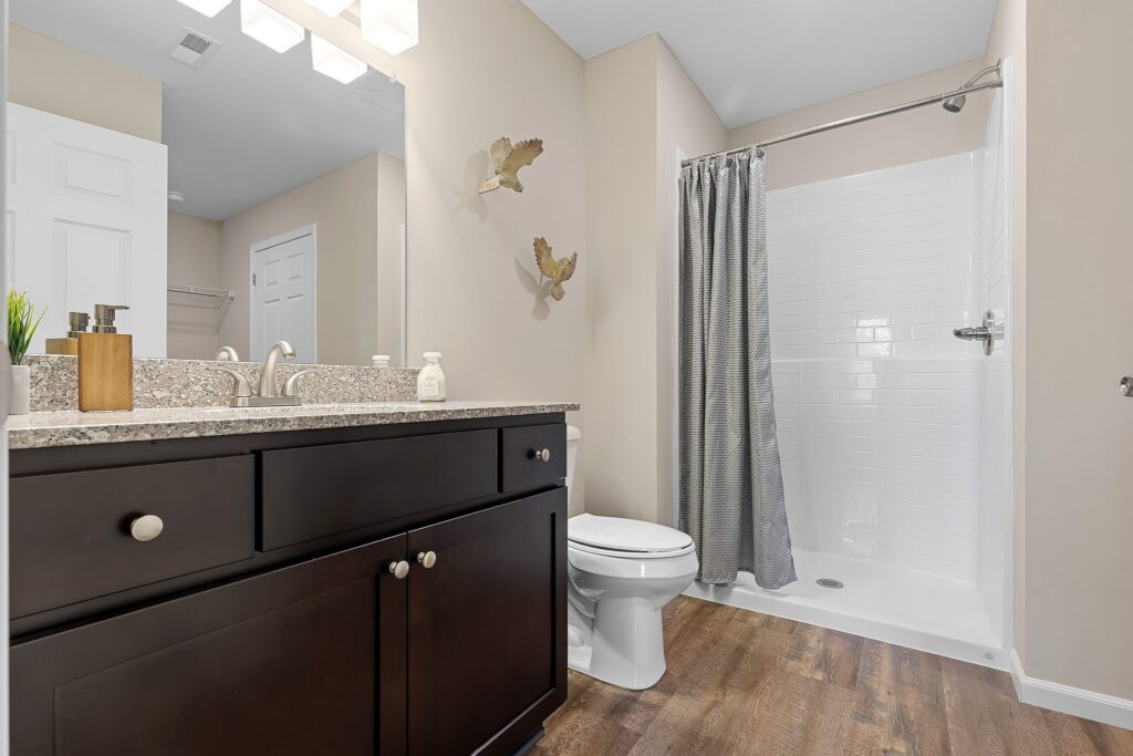 Modern bathroom with dark wood vanity, granite countertop, white toilet, walk-in shower with gray curtain, and wall decor above the toilet.