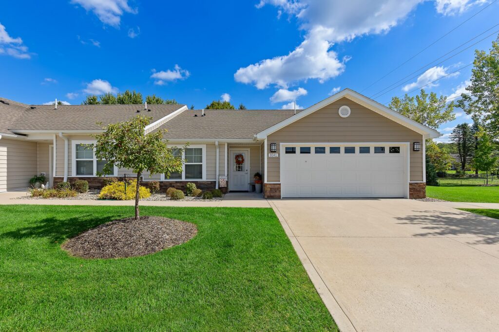 Single-story suburban house with beige siding, attached two-car garage, manicured lawn, small tree, and clear blue sky with scattered clouds.