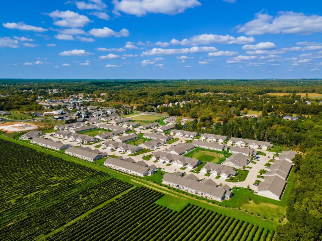 Aerial view of a suburban residential neighborhood bordered by farmland and forest under a blue sky with scattered clouds.