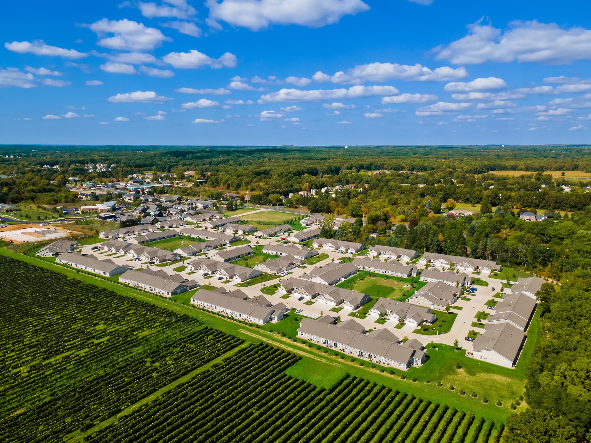 Aerial view of a suburban residential neighborhood bordered by farmland and forest under a blue sky with scattered clouds.