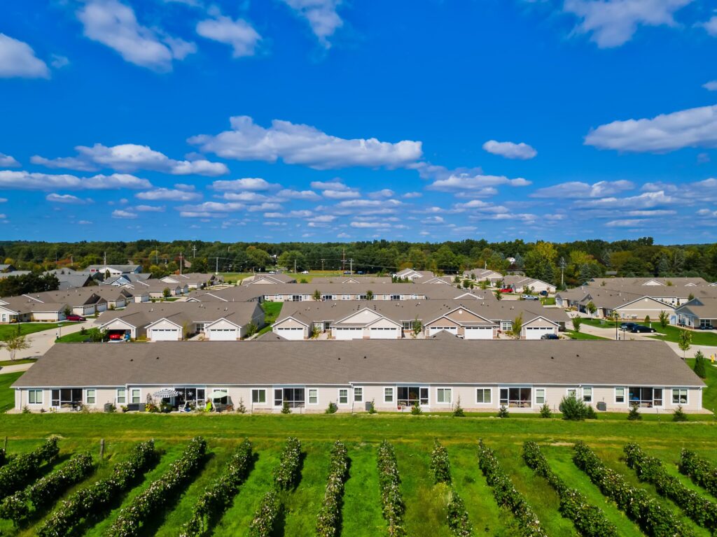 Aerial view of a residential neighborhood with single-story houses and a neatly arranged garden or vineyard in the foreground under a blue sky with scattered clouds.