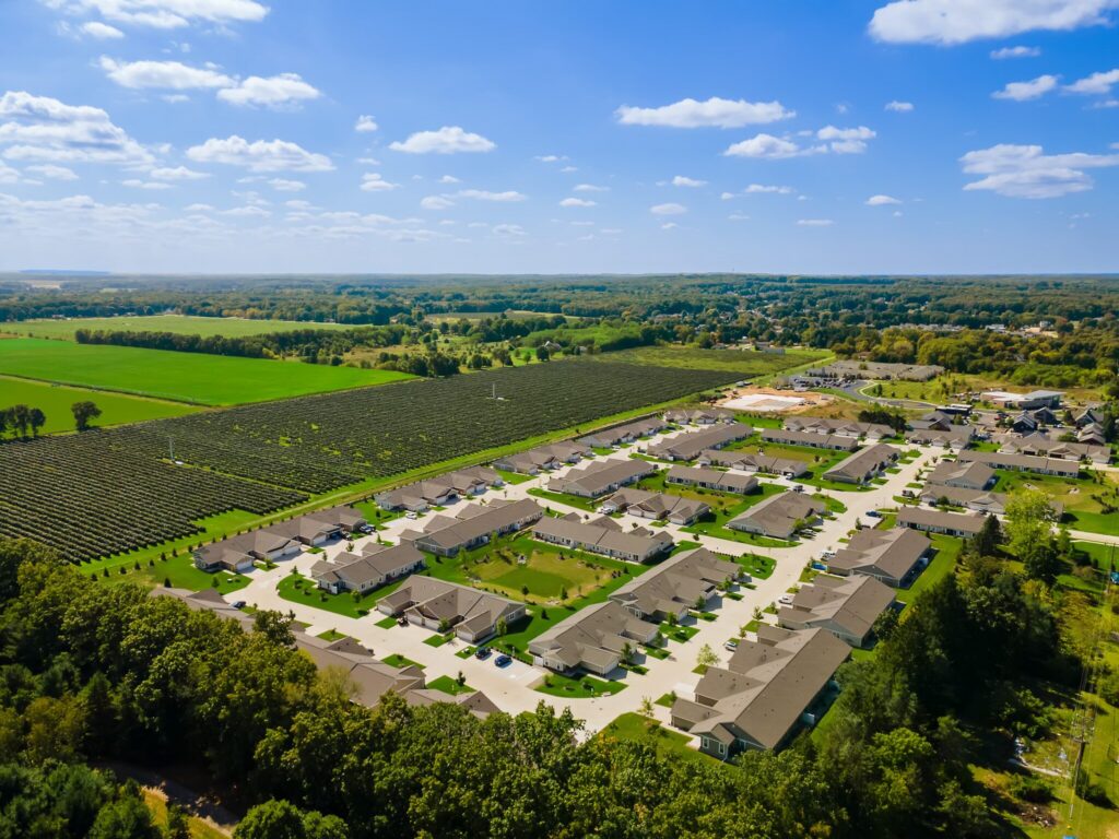 Aerial view of a suburban neighborhood with rows of houses, surrounded by green fields, trees, and blue sky with scattered clouds.
