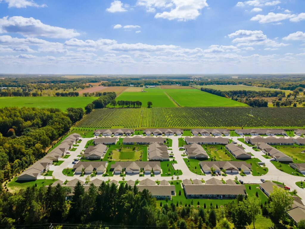Aerial view of a suburban neighborhood with rows of houses bordered by green fields and trees under a partly cloudy sky.