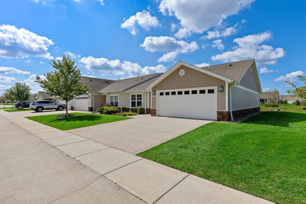Single-story suburban house with a double garage, light siding, green lawn, and a driveway under a blue sky with scattered clouds.