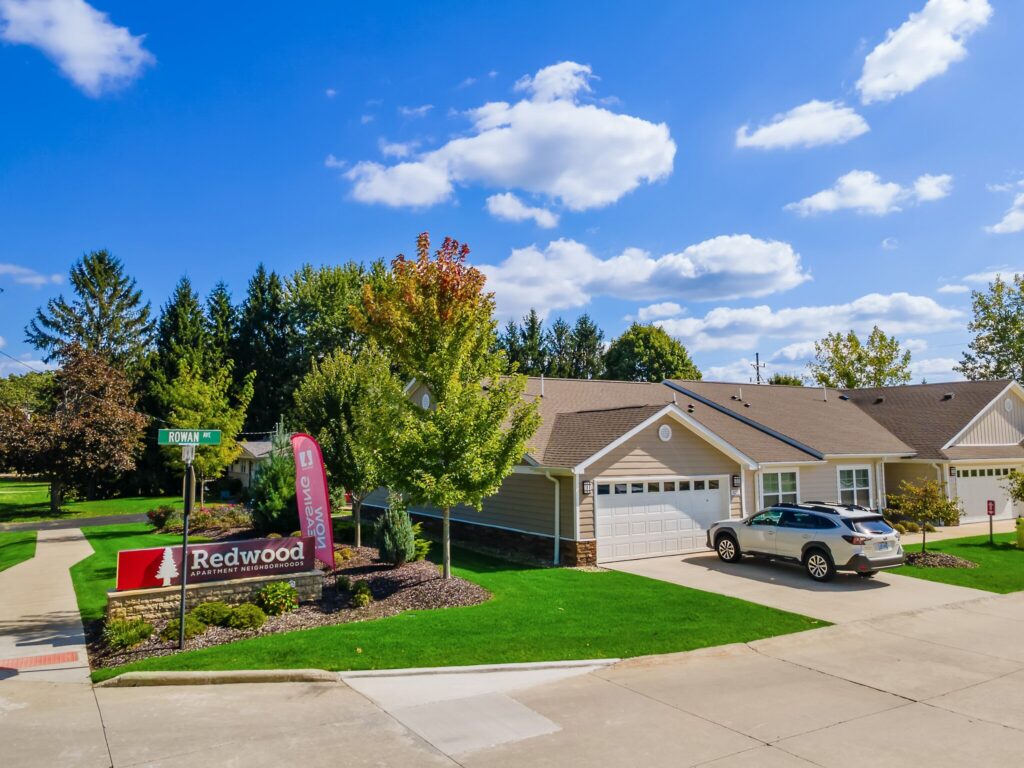 A suburban duplex with a two-car garage, a parked SUV, a "Redwood" community sign, and a street sign under a partly cloudy sky.