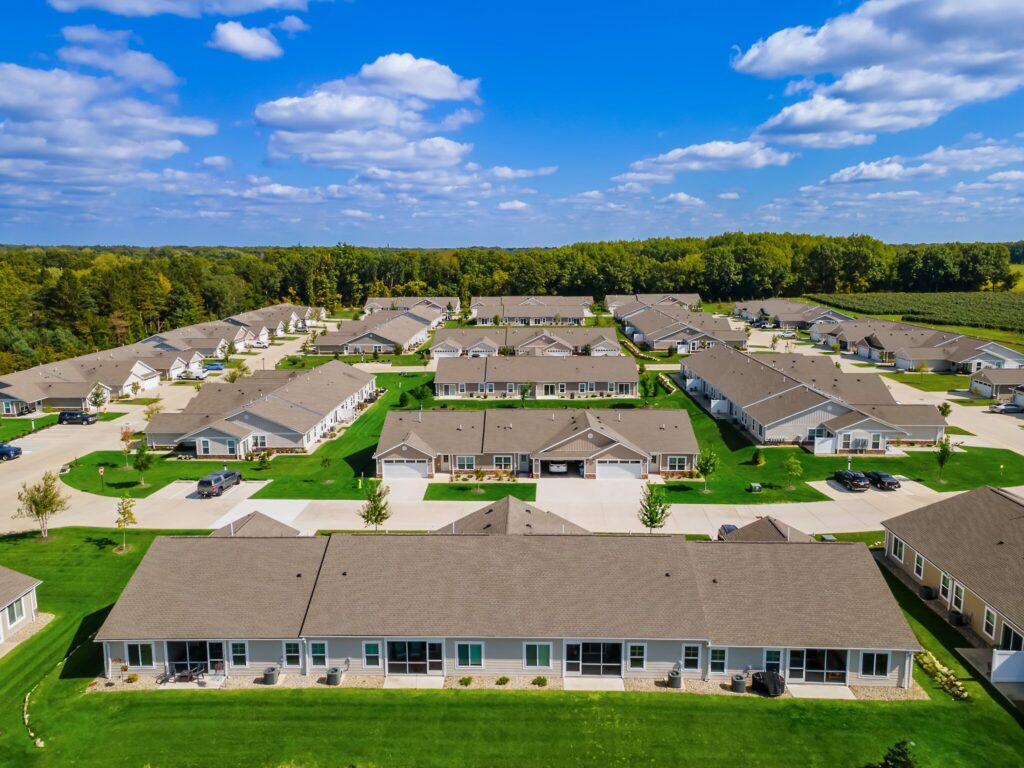 Aerial view of a suburban residential neighborhood with multiple single-story houses, green lawns, and tree-lined backgrounds under a blue sky with scattered clouds.