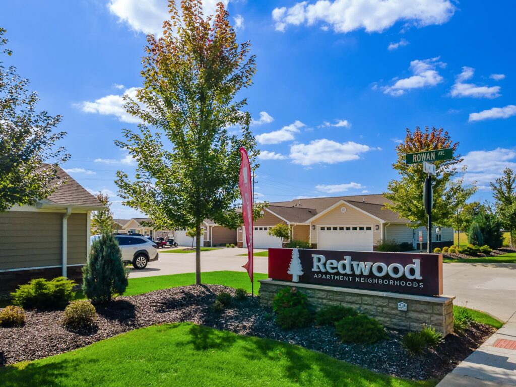 Entrance sign for Redwood Apartment Neighborhoods on a landscaped corner with houses, trees, a red flag, and a street sign for Rowan Ave under a blue sky.