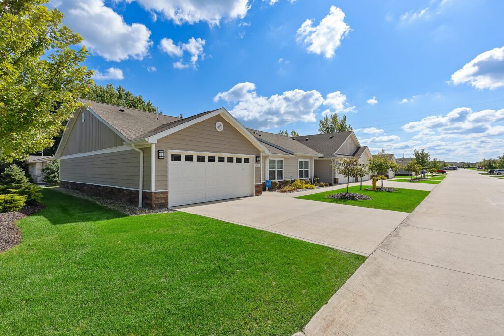 Single-story suburban house with a double garage, well-maintained lawn, and a wide driveway on a sunny day with partly cloudy skies.
