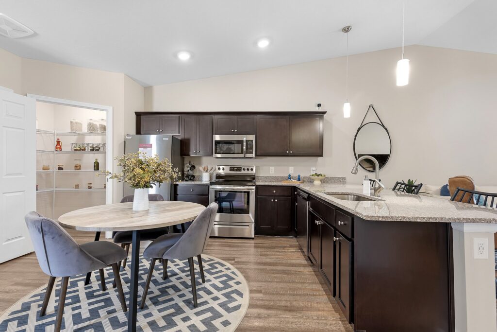 Modern kitchen with dark cabinets, stainless steel appliances, round dining table with two gray chairs, patterned rug, and open pantry in the background.