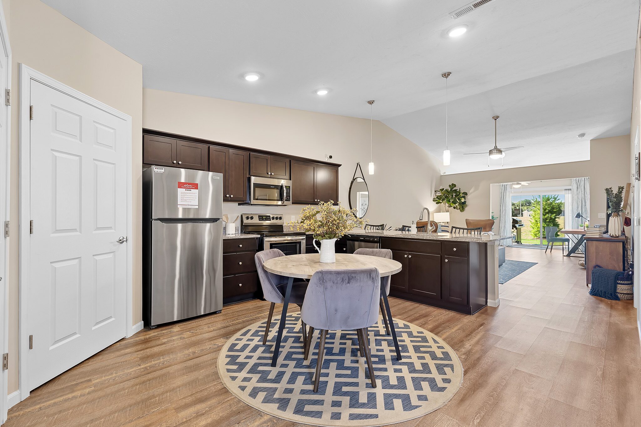 Open-concept kitchen and dining area with dark cabinets, stainless steel appliances, a round table with chairs on a patterned rug, and view of living room leading to a patio.