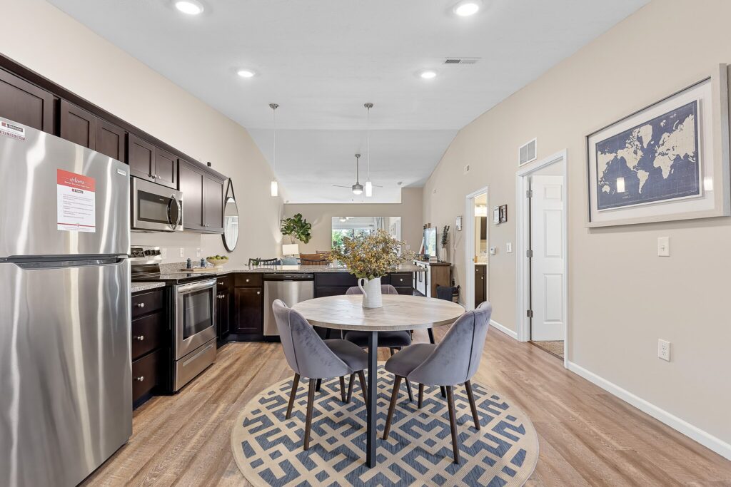 Modern kitchen and dining area with dark cabinets, stainless steel appliances, a round table with four gray chairs, light wood flooring, and wall art featuring a world map.
