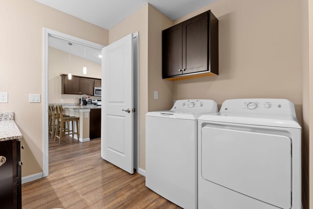 A laundry room with a white washer and dryer, dark wood cabinets, and a view into a kitchen with wood flooring and beige walls.