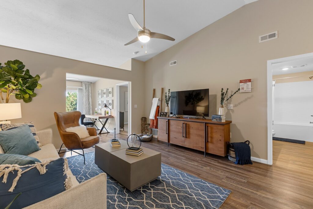 Modern living room with a sofa, armchair, TV on a wooden console, coffee table, and a rug. Open doorway leads to a workspace, and another to a bathroom. Ceiling fan and plant visible.