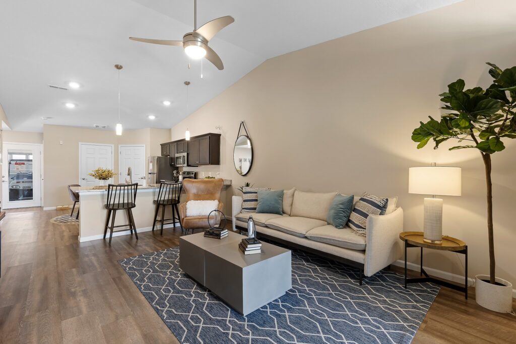 Modern open-plan living room and kitchen with neutral tones, a beige sofa, patterned rug, bar stools at the counter, and a potted plant in the corner.