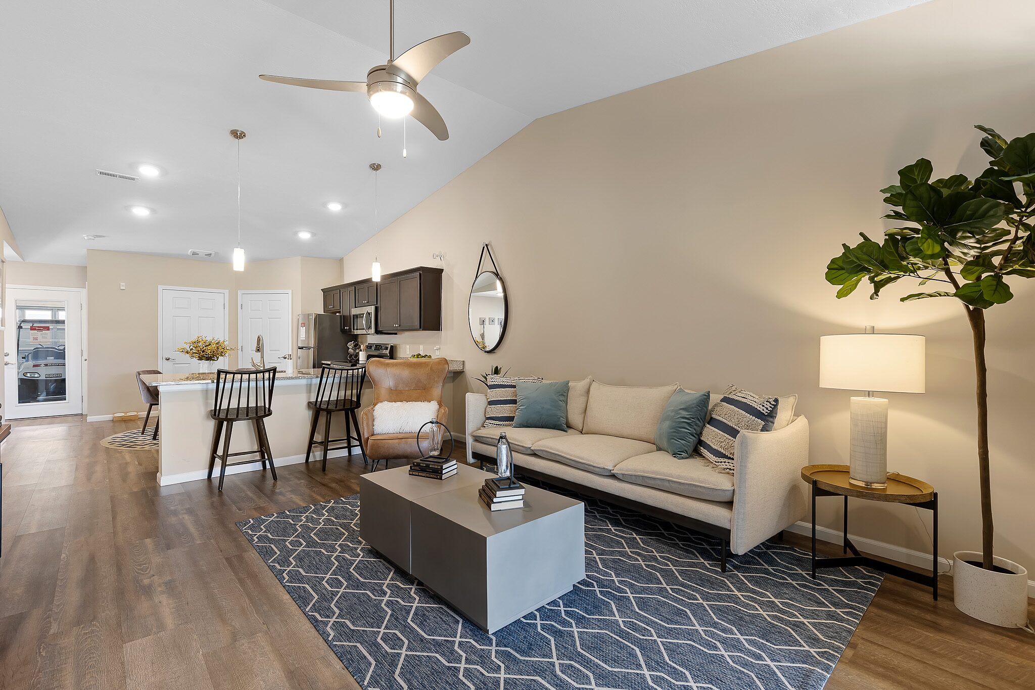 Modern open-plan living room and kitchen with neutral tones, a beige sofa, patterned rug, bar stools at the counter, and a potted plant in the corner.