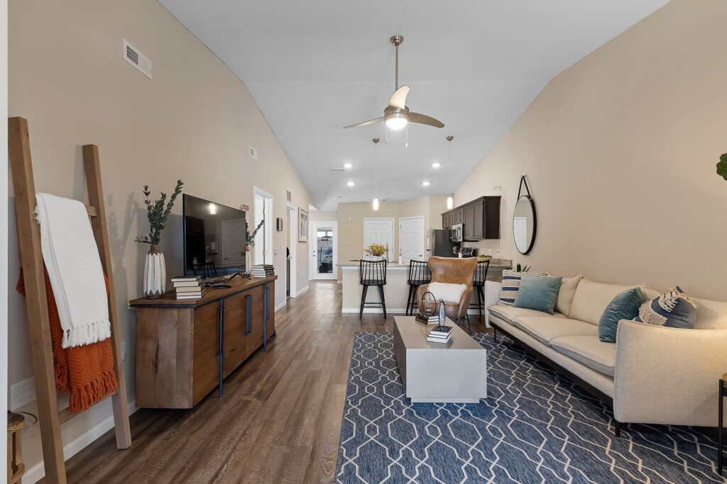 Modern living room with a beige couch, blue patterned rug, wooden floors, TV on a wooden console, dining area, and kitchen visible in the background.