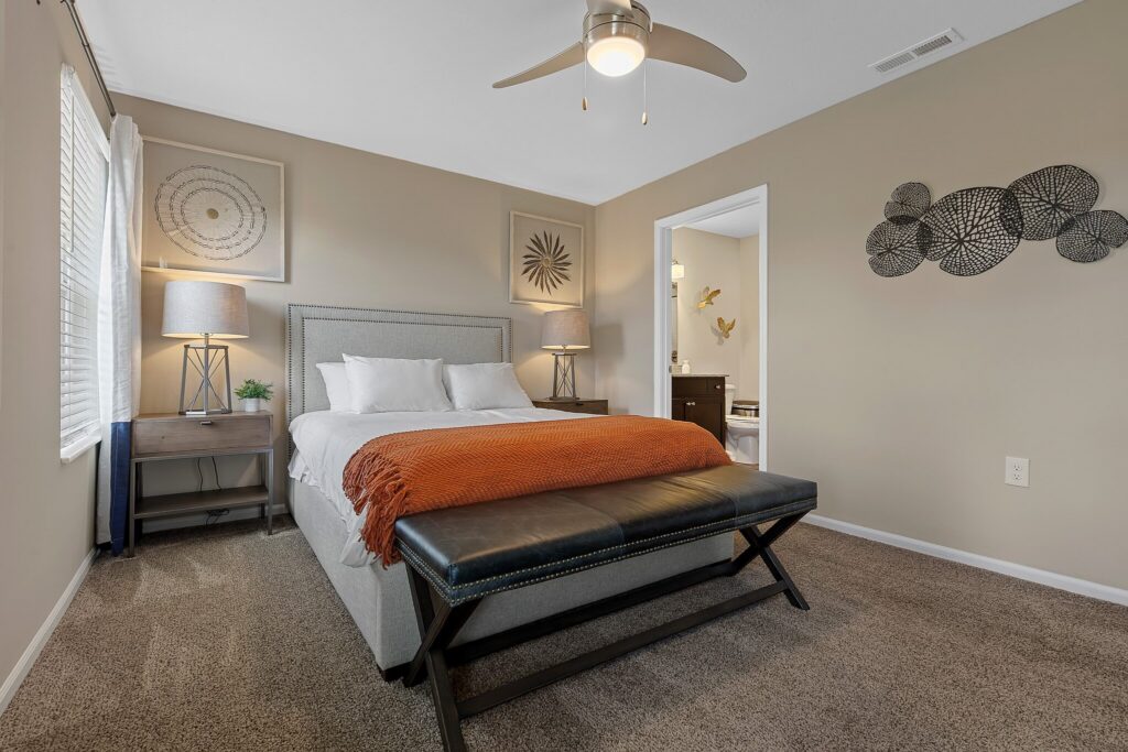 A neatly made bed with white linens and an orange throw sits in a carpeted bedroom with beige walls, a ceiling fan, two nightstands, and wall art.