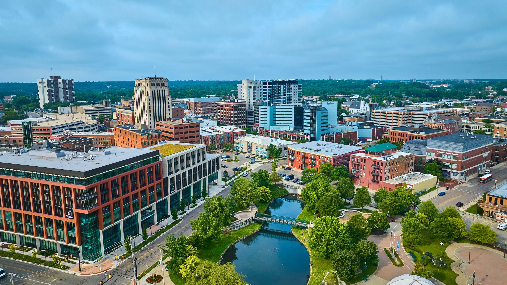 Aerial view of a downtown cityscape with modern and historic buildings, a small park, pond, and tree-lined streets under a cloudy sky.