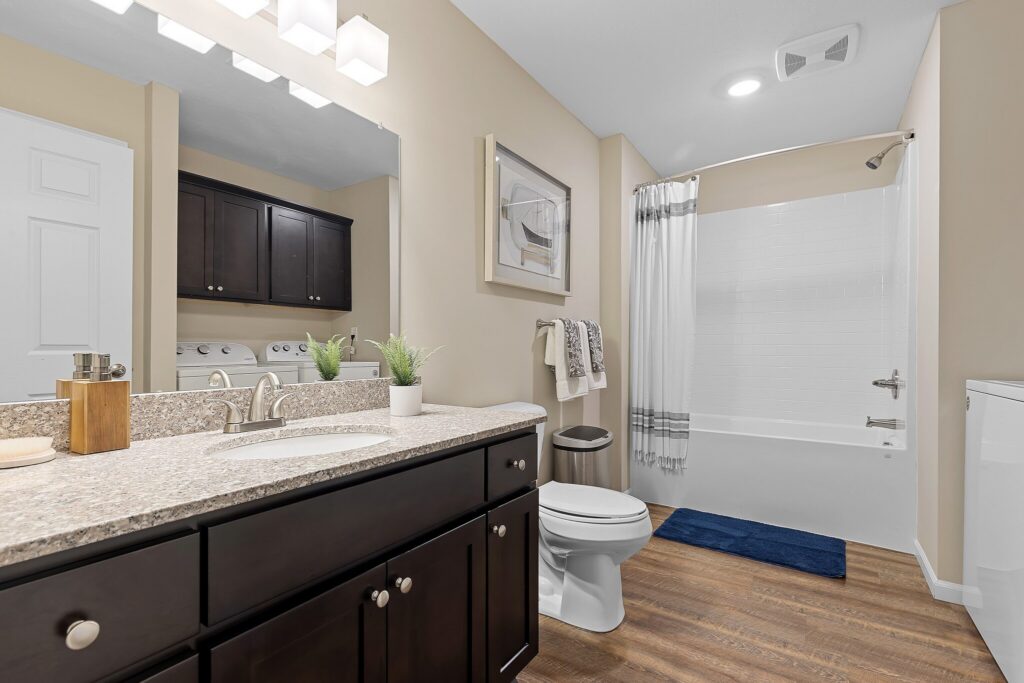 Modern bathroom with a granite countertop, dark wood cabinets, toilet, bathtub with a shower curtain, washer and dryer, and decorative plants and towels.