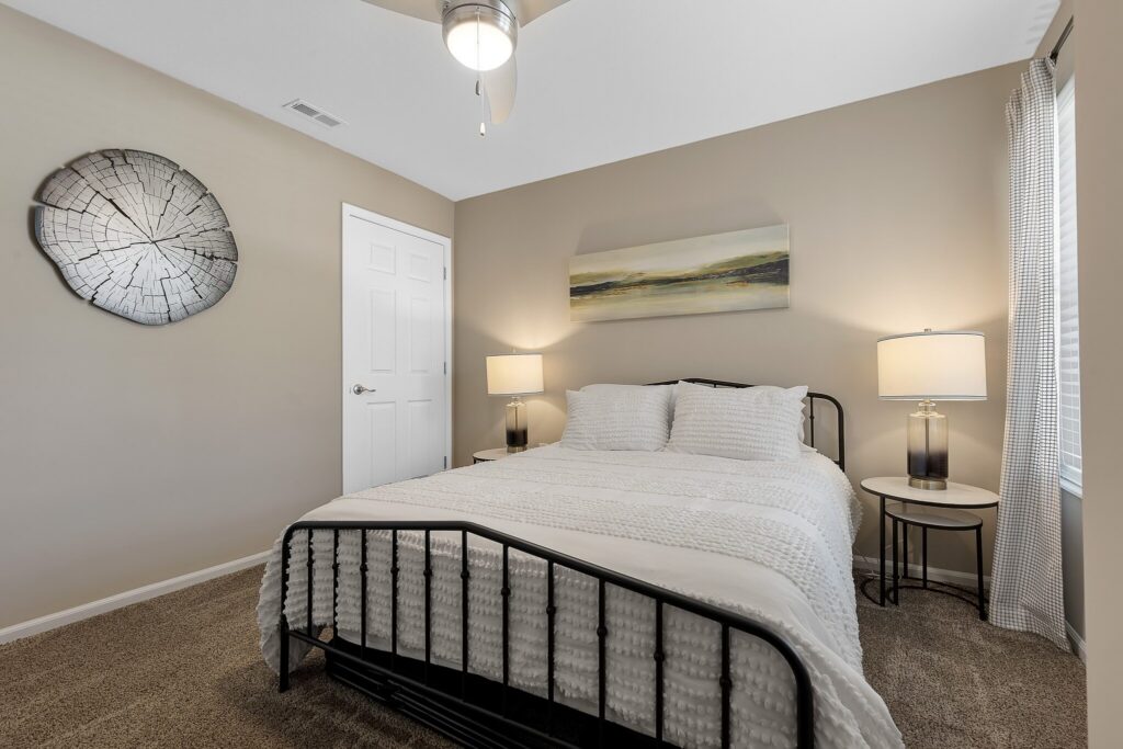 A bedroom with a metal bed frame, white bedding, two bedside tables with lamps, a circular wall art piece, and a landscape painting above the bed.