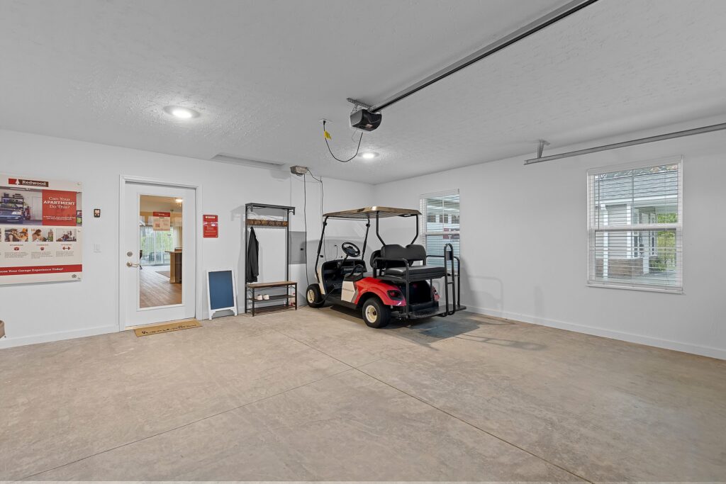 A spacious garage with a red golf cart parked inside, white walls, two windows, a door, and a few shelves and wall-mounted items.