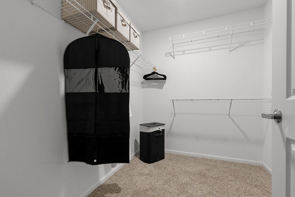 A mostly empty walk-in closet with wire shelves, a garment bag, a single hanger, three storage baskets, and a small black laundry hamper on carpeted flooring.