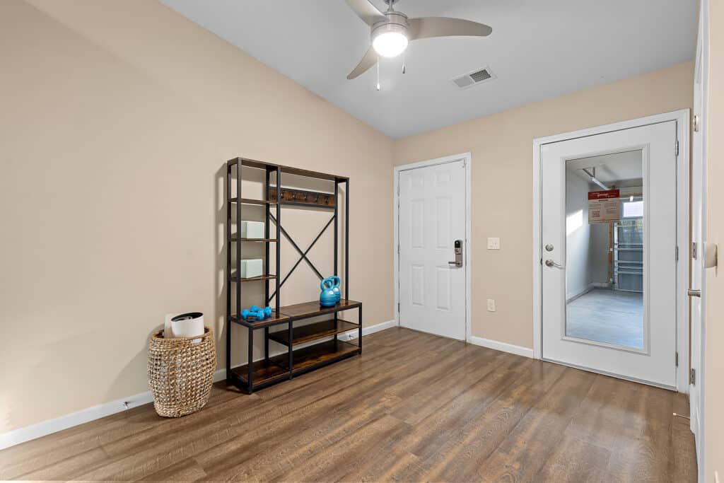A small, empty room with wood flooring, beige walls, a ceiling fan, a shelving unit, a wicker basket, and a door with a window leading to another room.