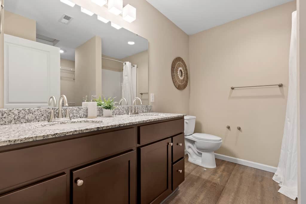 Modern bathroom with double sink vanity, granite countertop, wall-mounted mirror, toilet, towel rack, and wood-look flooring.