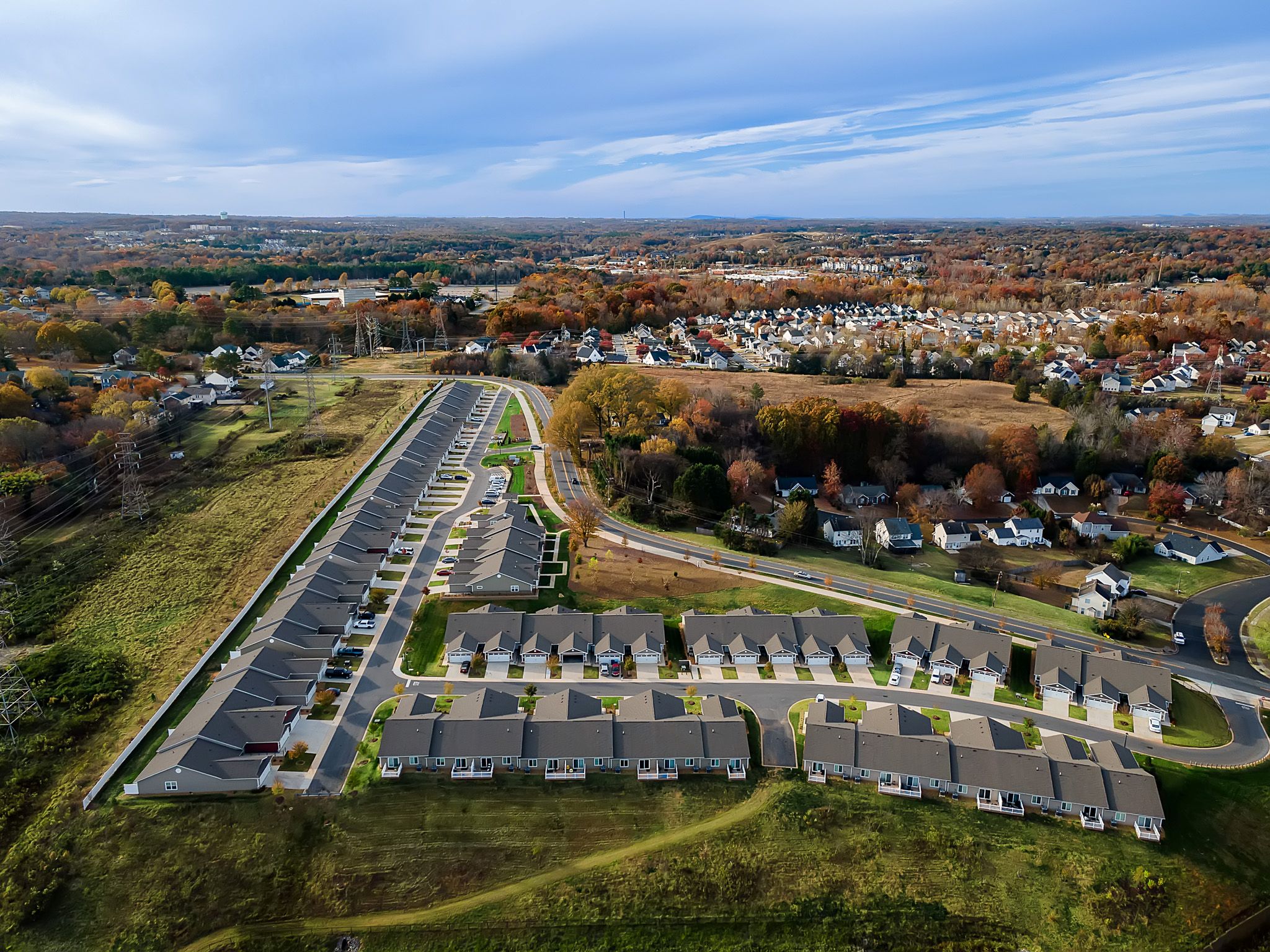 Aerial view of a suburban neighborhood with rows of houses, roads, grassy areas, and trees with autumn foliage stretching into the distance.