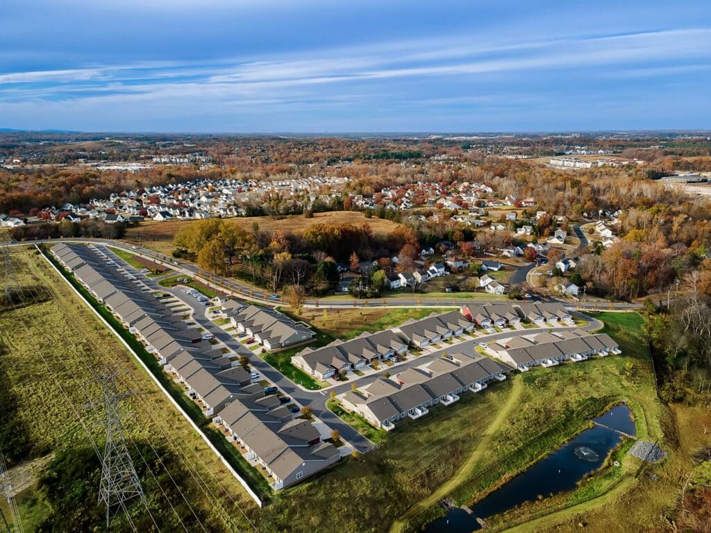 Aerial view of a suburban neighborhood with rows of houses, surrounding fields, a small pond, and distant tree-lined residential areas under a partly cloudy sky.