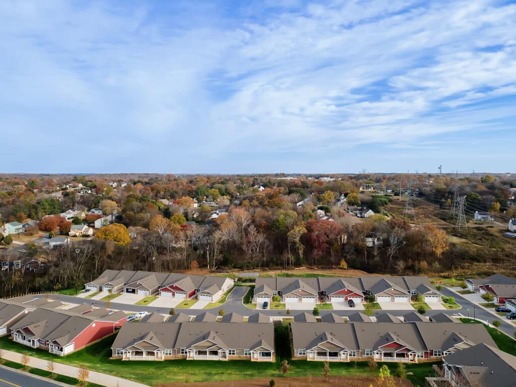 A suburban neighborhood with rows of single-story houses is shown in the foreground, while trees with autumn foliage and additional homes extend into the distance under a partly cloudy sky.