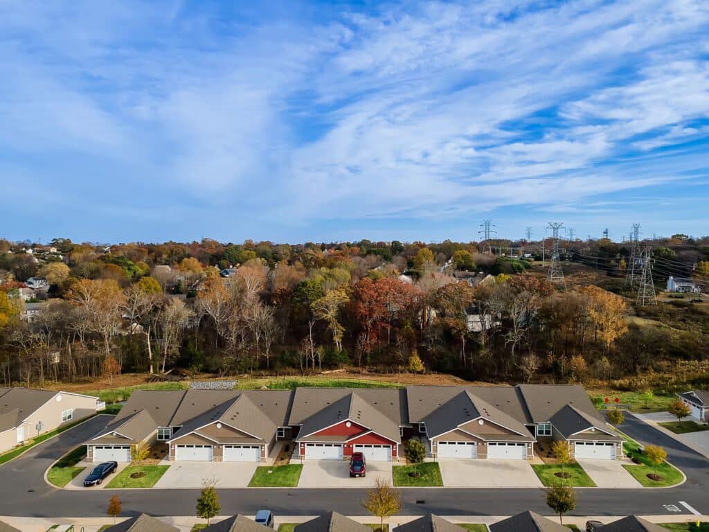 A row of suburban houses with garages faces a tree-lined area under a partly cloudy sky, with power lines and towers visible in the background.