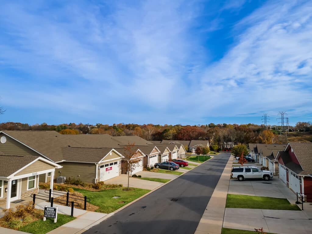 A suburban residential street with single-story houses, parked cars, green lawns, and autumn trees under a partly cloudy sky.
